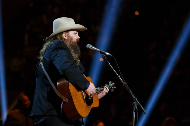 Chris Stapleton performs on stage at the CMA Music Festival in Nashville, Tennessee, playing guitar and singing into a microphone while wearing a hat, with a few people visible in the background.