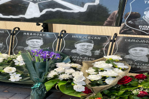 Ein Strauss bunter Blumen liegt auf einem Gehweg neben einem Schild, im Hintergrund sind Gedenktafeln zu sehen.