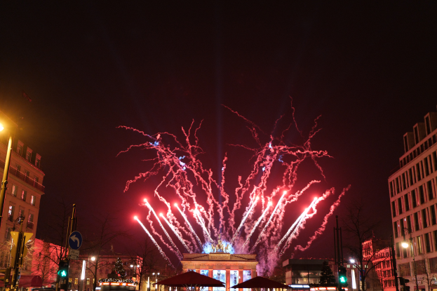 Eine belebte Stadtstraße in Berlin an Silvester, voller Menschen, Fahrzeuge und Gebäude, erhellt von Feuerwerk und Gebäudelichtern, die eine festliche Atmosphäre schaffen.