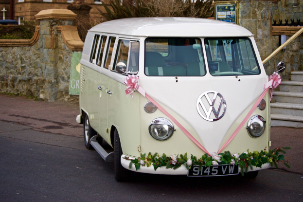 Ein weißer und pinkfarbener VW-Campervan mit floralen Verzierungen parkt vor einer Steinwand, rechts befinden sich Treppen und im Hintergrund ein Gebäude.