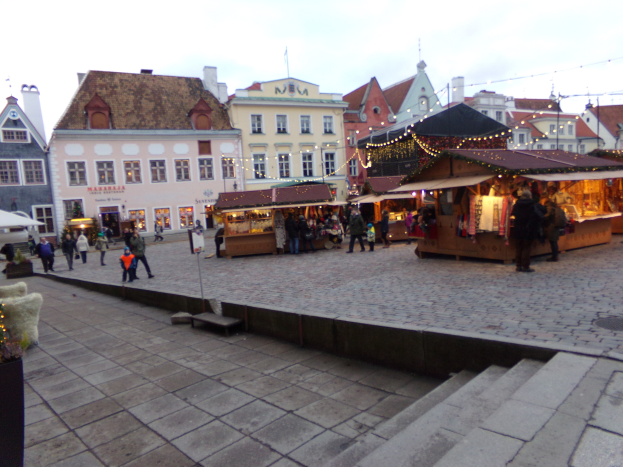 Ein geschäftiger Weihnachtsmarkt in Tallinn, Estland mit Menschen um geschmückte Stände, festlicher Beleuchtung, bewölktem Himmel, Treppen, die hinaufführen, und Topfpflanzen im Bereich.