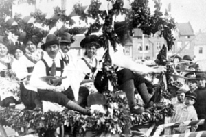 Schwarzes und weißes Foto von Menschen in traditioneller bayrischer Tracht auf einem geschmückten Umzugswagen, einige spielen Instrumente, mit Gebäuden und einem klaren Himmel im Hintergrund.