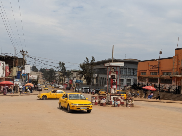 Ein gelbes Taxi fährt auf einer belebten Stadtstraße mit Gebäuden an den Seiten, während Fußgänger mit Regenschirmen auf dem Gehweg unter einem bewölkten Himmel gehen, mit Strommasten und Drähten im Hintergrund.
