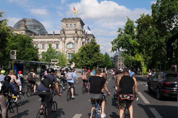 Eine Gruppe von Menschen fährt mit Fahrrädern eine von Bäumen gesäumte Straße in Berlin, Deutschland, hinunter, mit Gebäuden auf beiden Seiten, einem Bushaltestellen auf der rechten Seite und einer Flagge auf einem der Gebäude unter einem bewölkten Himmel.