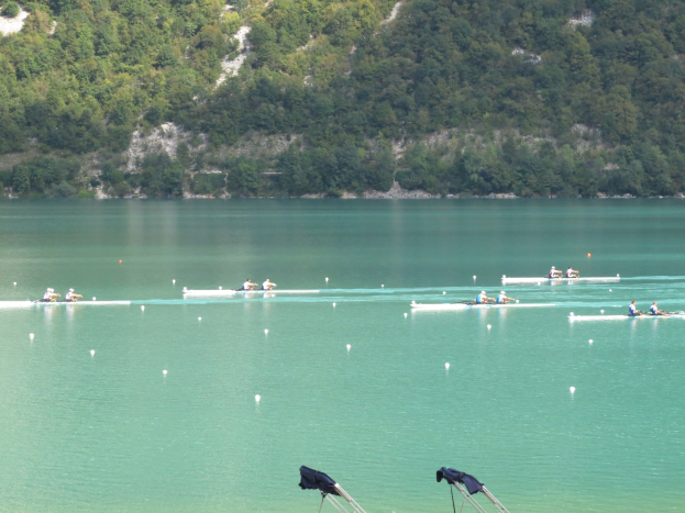 Gruppe von Menschen, die bei einem Wettbewerb auf einem Gewässer mit Bäumen und einem Hügel im Hintergrund rudern, mit Kleiderständern am Boden.