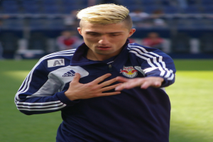 Ein junger Mann in einem blauen T-Shirt steht auf einem Fußballfeld und streckt die Arme aus, mit entschlossener Miene und einem unscharfen Hintergrund.