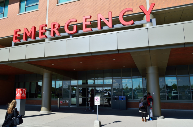 Eine Gruppe von Menschen geht vor einem Glaswand-Notfallgebäude mit Säulen, einem Namensschild, Hinweistafeln auf Pfählen und einem Müllcontainer.