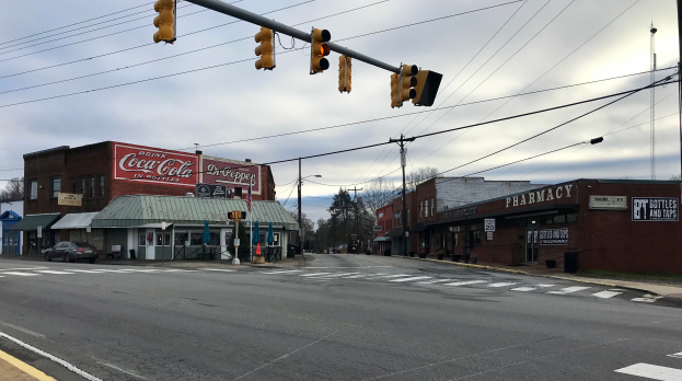Eine Straßenkreuzung mit Verkehrsampel, Fahrzeugen, Gebäuden mit Namensschildern, Strommasten mit Drähten, Bäumen und einem bewölkten Himmel.