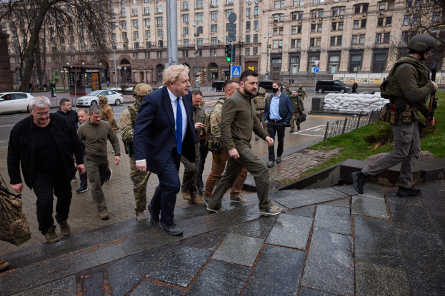 Eine Gruppe von Menschen, einschließlich Boris Johnson, die eine Straße entlanggehen, neben einem Gebäude, wobei einige Helme tragen und Waffen halten, mit Fahrzeugen, Laternenmasten, Verkehrsampeln, Bäumen und Gebäuden im Hintergrund.