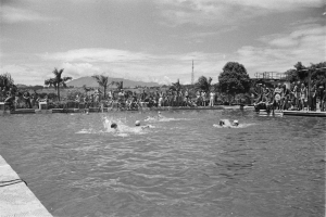 Schwarze und weiße Fotografie von Menschen, die in einem Pool schwimmen, mit Zuschauern hinter einem Zaun und Bäumen und einem bewölkten Himmel.