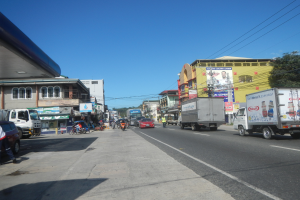 Eine belebte Straße in der Stadt mit zahlreichen Autos, Lastwagen, Füpßen, Gebäuden, Schildern, Strommasten, Bäumen und einem bewölktem Himmel.
