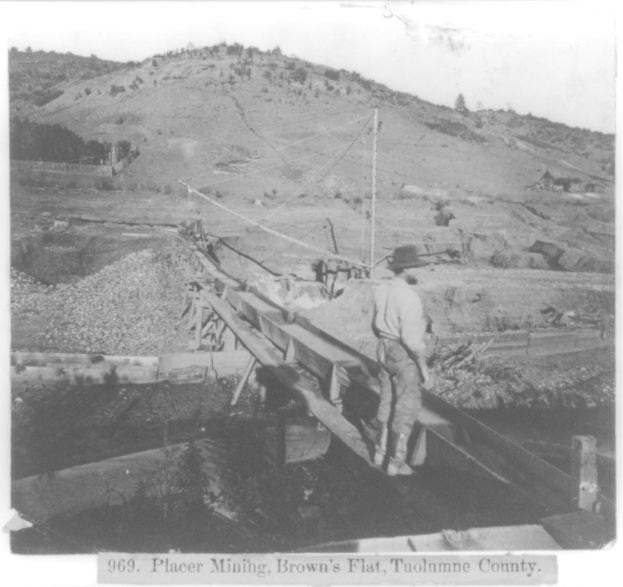 Schwarzes und weißes Foto eines Mannes mit Hut auf einer hölzernen Brücke in Placer Mining, Brown's Flat, Tuolumne County, umgeben von Pfählen, Bäumen und Hügeln mit sichtbarem Himmel und Text unten.