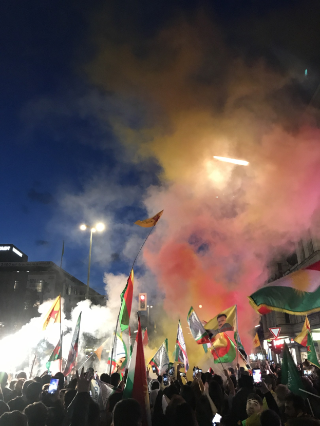 Menschenmenge vor einem Gebäude mit Fahnen schwenkend und Handys haltend unter farbigen Feuerwerkskörpern