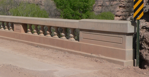 Eine Betonbrücke überspannt einen Schotterweg, mit einem gelben und schwarzen Schild darauf, umgeben von Bäumen und Felsen.