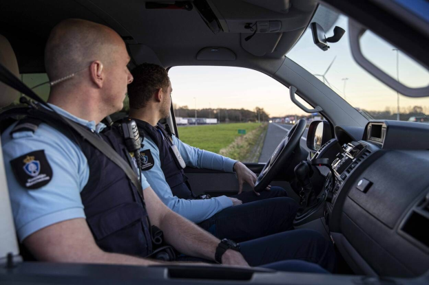 Zwei Polizisten in einem Auto auf einer Straße mit Gras, Bäumen und Polen im Hintergrund unter einem klaren blauen Himmel.