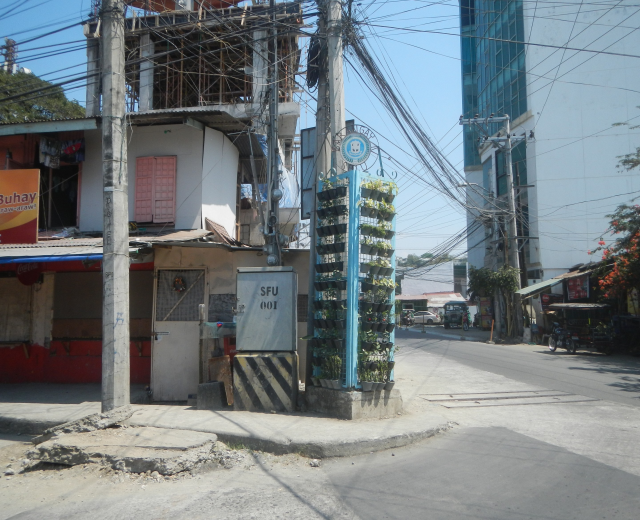 Eine Stadtstraße mit Gebäuden, Strommasten, Kabeln, Schildern, Fahrzeugen, Bäumen und Himmel, mit einem Gebäude mit Drähten an der Seite im Vordergrund.