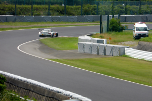 Ein Rennwagen fährt eine Rennstrecke entlang, im Hintergrund ist ein Polizeiauto zu sehen, umgeben von Gras, Pflanzen, Bäumen und einem Zaun, mit einem Fahrzeug auf der rechten Seite und Text unten.