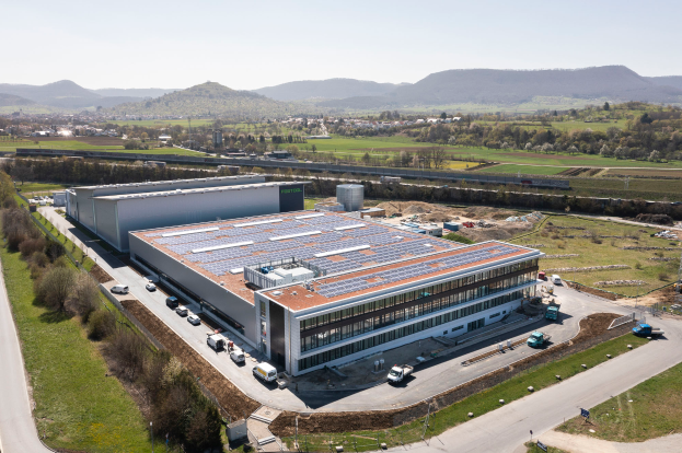 Eine Luftaufnahme eines großen Fabrikgebäudes mit Solarmodulen auf dem Dach, umgeben von Fahrzeugen, Bäumen und Masten, mit Hügeln und einem klaren blauen Himmel im Hintergrund, als erstes solarbetriebenes Werk in Deutschland vermerkt.