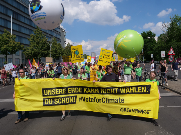 Eine Gruppe von Menschen hält ein gelbes 'Stimme für den Klimaschutz'-Plakat, während sie eine Straße entlanggehen, mit Luftballons, Schildern, Fahrrädern, Bäumen, Laternenpfählen und Gebäuden im Hintergrund unter einem klaren blauen Himmel.