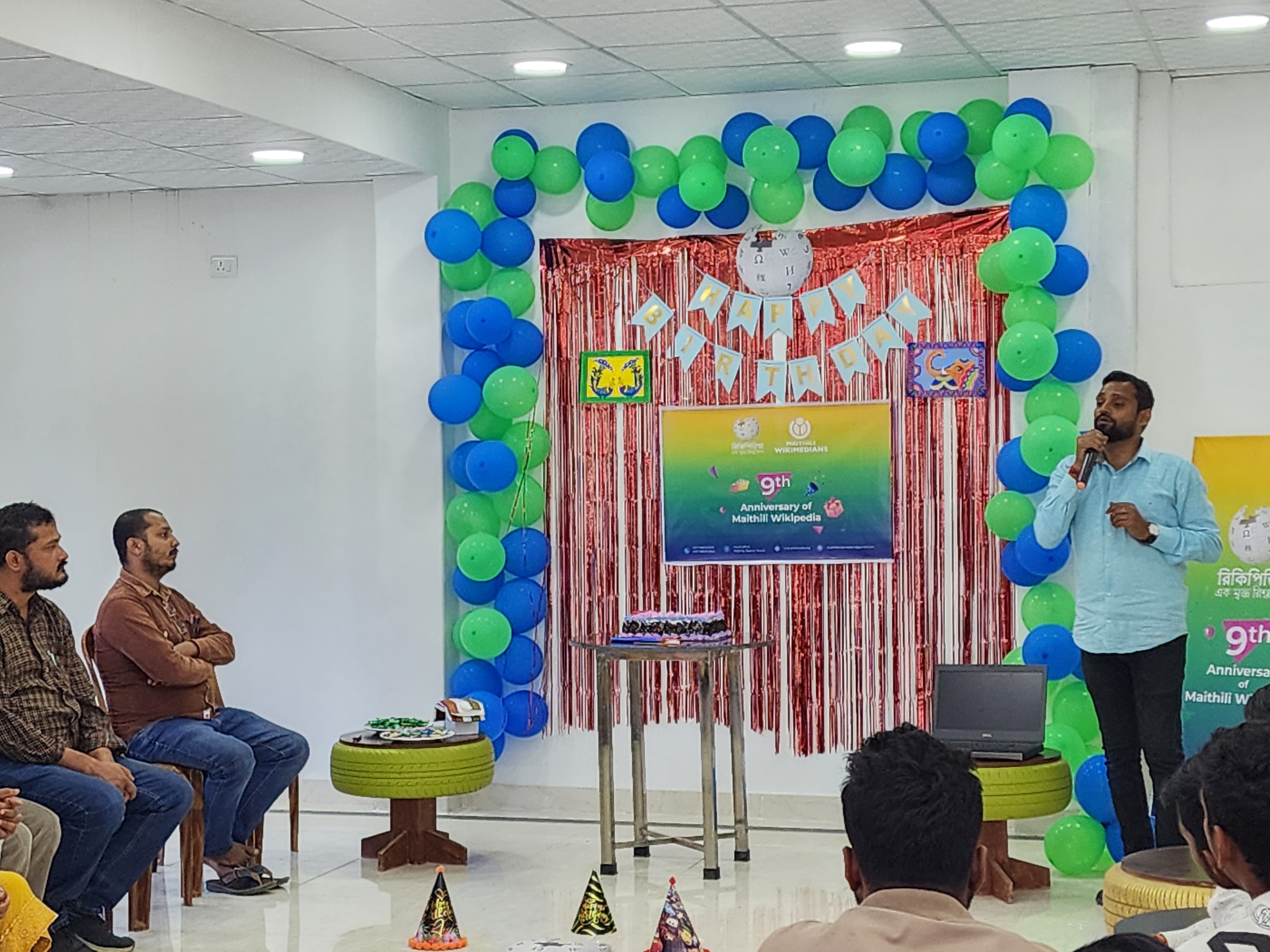 Eine Gruppe von Menschen, die auf Stühlen sitzen und einer dekorierten Bühne mit Luftballons, einem Banner, einer Uhr und anderen Gegenständen zugewandt sind, mit einem Tisch mit einem Laptop und einer Person, die ein Mikrofon hält, während einer 9. Geburtstagsfeier.