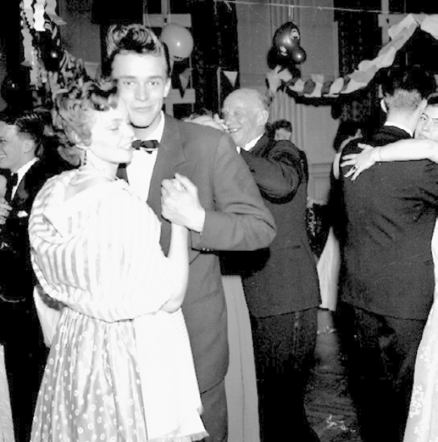 Ein Schwarz-Weiß-Foto von einem Mann und einer Frau, die auf einer Party tanzen, umgeben von lachenden Menschen, mit Ballons und Dekorationen im Hintergrund.