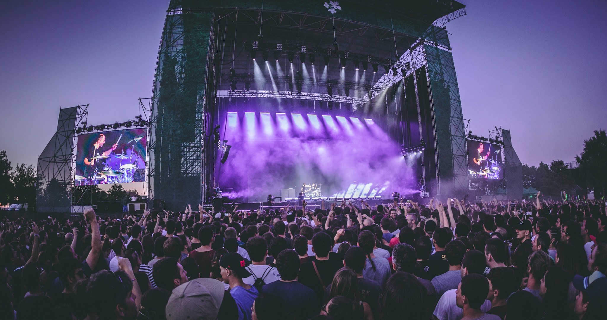 Eine große Menschenmenge bei einem Musikfestival schaut auf einer beleuchteten Bühne mit Instrumenten und Bildschirmen, die vor einem Gebäude, Bäumen und einem klaren blauen Himmel aufgebaut sind.