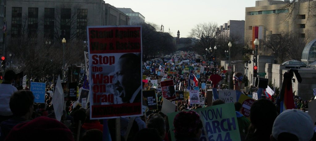 Menschen protestieren auf einer Straße und halten Schilder hoch, mit Gebäuden, Bäumen, Laternen und dem Himmel im Hintergrund.