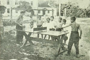 Eine Gruppe von Kindern spielt Tischtennis vor einem Gebäude mit Fenstern, umgeben von Bäumen und Pfosten, in einem Schwarz-Weiß-Bild mit Text unten.