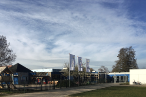 Ein gro├če Geb├Ąude mit einer gr├╝nen Fl├Ąche davor, umgeben von einem Metallzaun, einem Weg, B├Ąumen, Pfosten, Bannern, einem geparkten Fahrrad, n├Ąheren H├Ąusern und einem bew├Âlktem Himmel, das als neuer Spielplatz f├╝r Kinder dient.