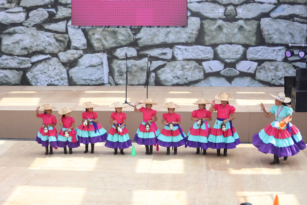 Gruppe junger Mädchen in bunten Kleidern und Hüten, die auf einer Bühne mit Verkehrskegeln, Mikrofonen, Lautsprechern, Scheinwerfern und einem Bildschirm mit Text im Hintergrund tanzen, während das Publikum von unten zusieht.