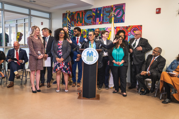 Gruppe von Menschen vor einem Rednerpult mit Mikrofon und einer Tafel mit Text, einige sitzen auf Stühlen und andere stehen, mit Flaggen, Feuerlöscher und Deckenleuchten im Hintergrund.