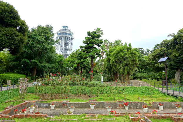 Ein Park mit einem Turm im Hintergrund, umgeben von grünem Gras, Pflanzen, Bäumen und Blumentöpfen, mit einer Straße mit Geländern, einem Pfahl mit einer Tafel und einem sichtbaren Himmel über dem Turm.