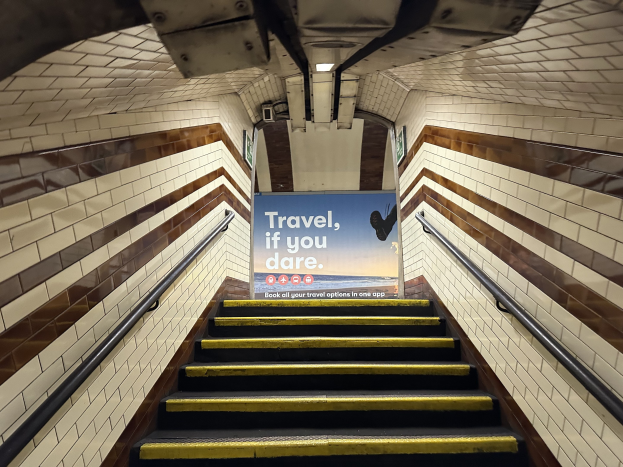 Eine Treppe in einer U-Bahn-Station mit einem "Reise, wenn du wagst"-Schild oben, flankiert von Geländern und sichtbaren Stationswänden im Hintergrund.
