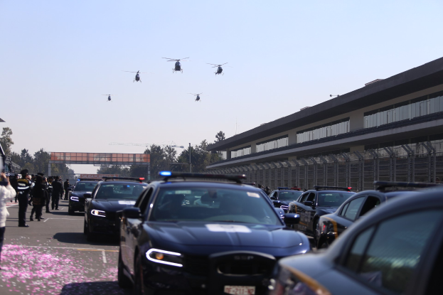 Eine Gruppe von Polizeiwagen vor einem Gebäude geparkt, mit Menschen auf der linken Straßenseite und vier Hubschraubern in der Luft, mit Bäumen, Pfählen, einer Brücke und einem klaren blauen Himmel im Hintergrund.