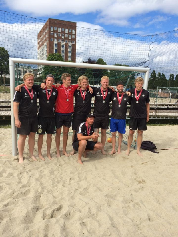 Mannergruppe in Sportbekleidung auf einem sandigen Strand, mit Medaillen und einem Netz umgeben, mit Bäumen, Gebäuden und einem bewölkten Himmel im Hintergrund.