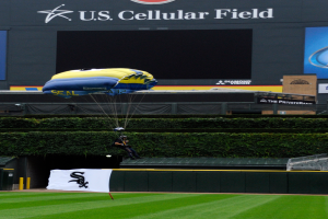 Eine Person gleitet mit einem Gleitschirm über ein Baseballfeld mit einem Fallschirm, umgeben von saftigem Grün, Pflanzen und einem Banner mit Text, mit Tafeln mit Text und Logos und einem Gebäude im Hintergrund.