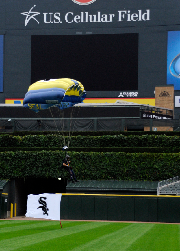 Eine Person gleitet mit einem Gleitschirm über ein Baseballfeld mit einem Fallschirm, umgeben von saftigem Grün, Pflanzen und einem Banner mit Text, mit Tafeln mit Text und Logos und einem Gebäude im Hintergrund.