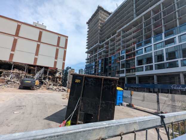 Großes Gebäude mit Schutt davor, umgeben von einem Metallzaun, einem Container, Pflanzen, Bäumen, Pfählen, einem Schild, Fahrzeugen, Steinen und einem bewölkten Himmel auf einer Baustelle für ein neues Wohngebäude.