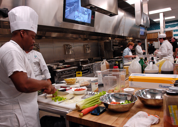 Eine Gruppe von Köchen in einer Küche beim Zubereiten von Speisen auf einem Schneidbrett, wobei ein Koch in der foreground in einer weißen Schürze und einer Mütze steht und ein Messer hält und von Utensilien, Schüsseln, Gemüse, einem Mobiltelefon, Flaschen und anderen Gegenständen umgeben ist, mit einem Fernseher und Deckenlampen im Hintergrund.