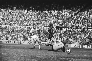 Schwarzes und weißes Foto von Männern, die auf einem Feld Fußball spielen, mit Zuschauern im Hintergrund und einer 'Bundesliga 1978-79 Ajax 1-1 Hannover 96'-Fahne.