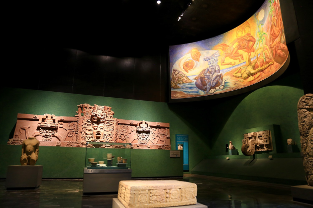 Ein Museumsraum mit verschiedenen Artefakten, darunter Skulpturen, Tischobjekte, eine Texttafel, eine Wandmalerei und Deckenleuchten im Anthropologischen Museum von Mexiko-Stadt.