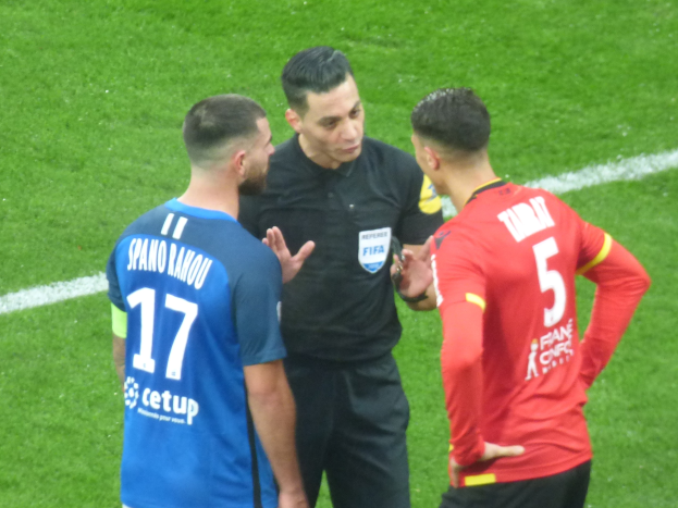 Ein Schiedsrichter in schwarzer Kleidung spricht mit zwei Männern auf einem Fuballfeld, einer in blauen und roten Shirts mit Text, beide tragen Brillen.