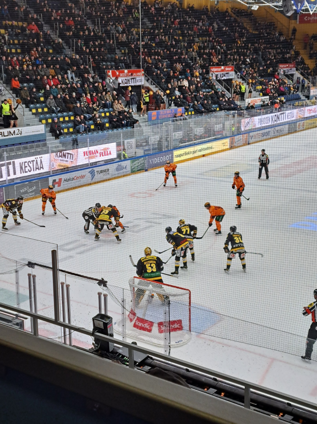 Eine Gruppe von Menschen, die Hockey auf einem Eisring mit einem Netz und einer umliegenden Absperrung spielen, Zuschauer sitzen im Hintergrund und Banner mit Text, unter Stadionbeleuchtung.