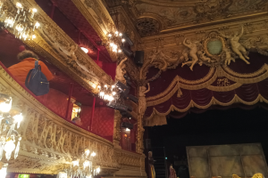 Das Innere eines prunkvoll dekorierten Theaters mit Kronleuchtern an der Decke, Skulpturen an den Wänden und einigen Menschen auf der Bühne, mit verstreuten Gegenständen, die eine lebendige Atmosphäre schaffen.