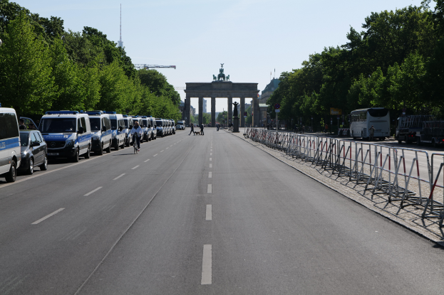 Eine lange Reihe von Polizeiwagen steht auf der Seite einer Straße vor dem Brandenburger Tor in Berlin, Deutschland, mit Menschen, die Fahrräder fahren und auf der Straße stehen, sowie Absperrungen und Bäume an den Seiten und einem Bogen mit Statuen im Hintergrund.