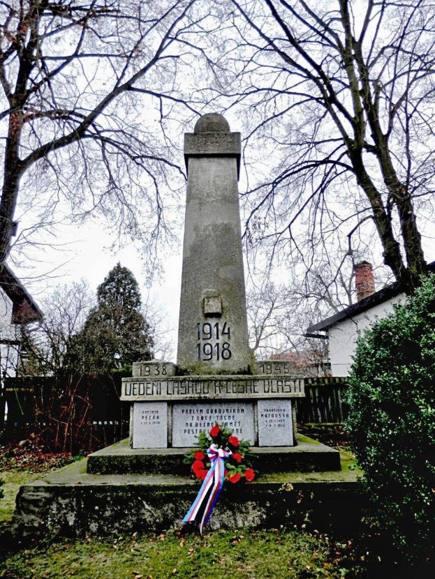 Ein Weltkriegsgedenkmal in einem Friedhof, geschmückt mit einem Kränzchen, umgeben von Gras und welken Blättern, mit Pflanzen, Bäumen, Häusern und Himmel im Hintergrund.