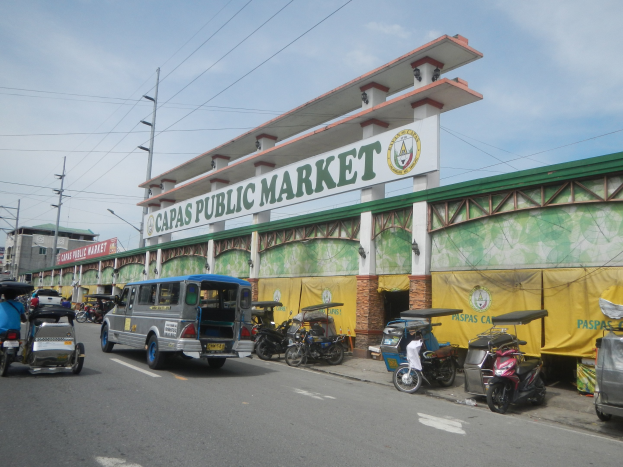 Eine belebte Stadtstraße mit Autos, Motorrädern und Rikschas vor einem Gebäude mit einer "Capas Public Market"- Tafel, Strommasten, Laternenmasten und bewölktem Himmel im Hintergrund.