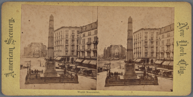 Altes Schwarz-Weiß-Foto einer Stadtstraße mit einem zentralen Denkmal, umgeben von Gebäuden, Straßeneinrichtungen, parkenden und fahrenden Fahrzeugen, Fußgängern auf dem Gehweg und einem sichtbaren Himmel.