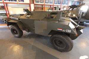 Ein gepanzertes Fahrzeug steht in einem Gebäude geparkt, umgeben von anderen Fahrzeugen, mit Texttafeln, Regalen, einer Tür und Deckenleuchten im Hintergrund.