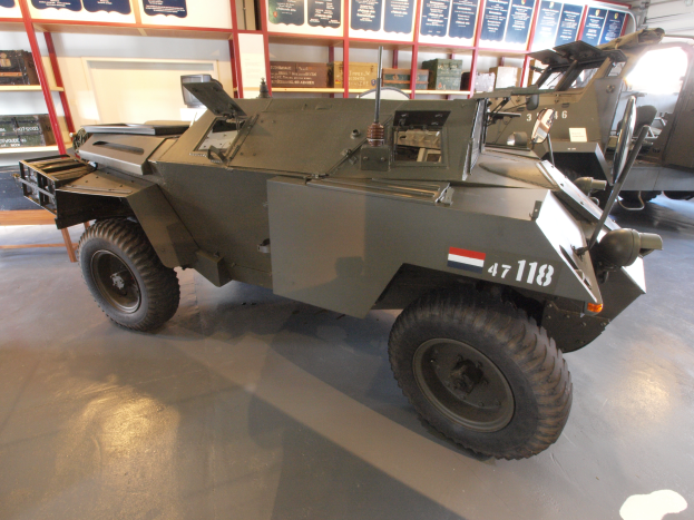 Ein gepanzertes Fahrzeug steht in einem Gebäude geparkt, umgeben von anderen Fahrzeugen, mit Texttafeln, Regalen, einer Tür und Deckenleuchten im Hintergrund.